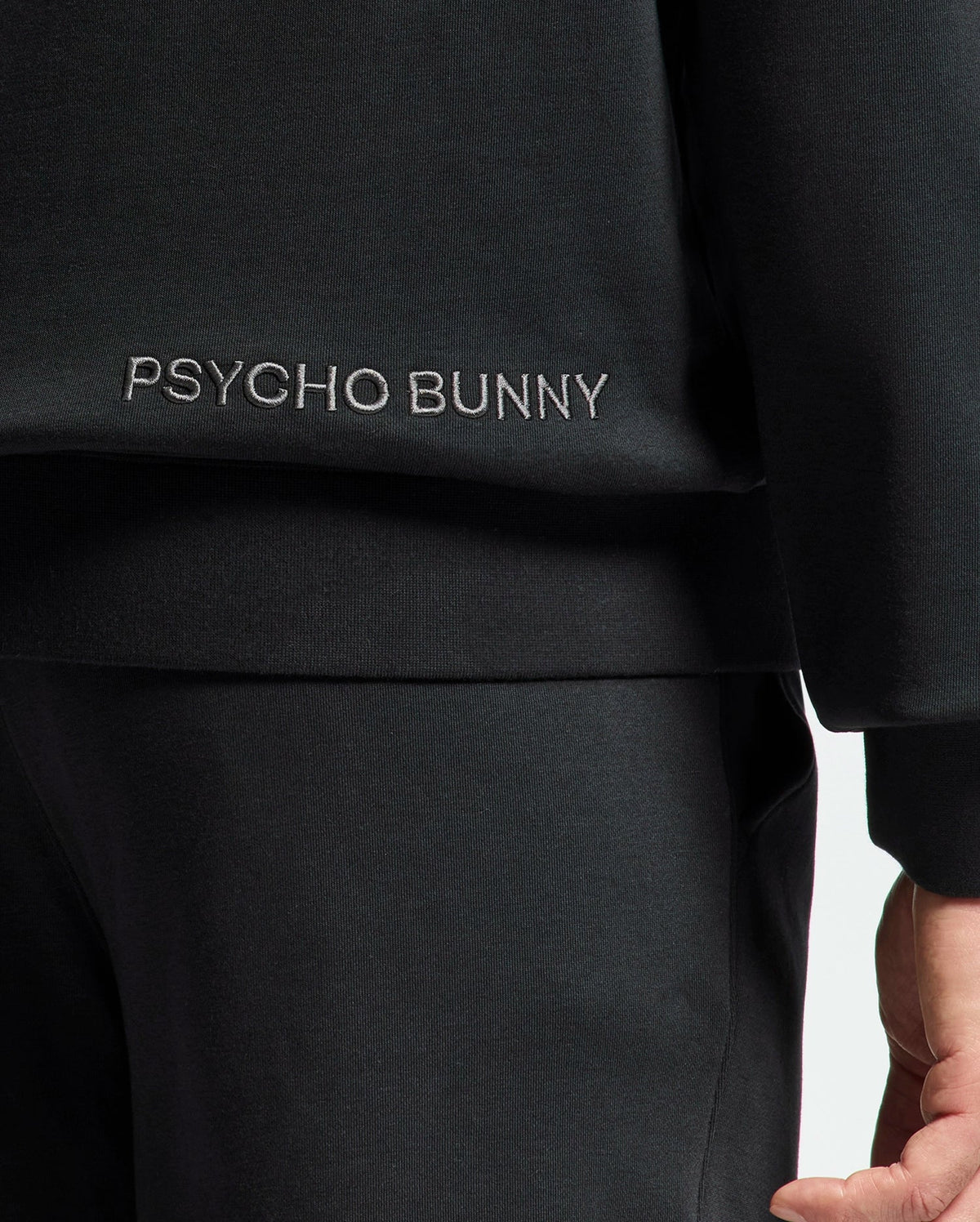Close-up of a person in a black MENS NEW CASTLE FRENCH TERRY HOODIE by Psycho Bunny. The sleeve, torso, and pants are visible. "Psycho Bunny" is embroidered in white on the side, adding elegance. The person's hand is slightly showing.
