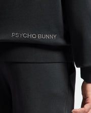 Close-up of a person in a black MENS NEW CASTLE FRENCH TERRY HOODIE by Psycho Bunny. The sleeve, torso, and pants are visible. "Psycho Bunny" is embroidered in white on the side, adding elegance. The person's hand is slightly showing.