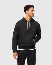 A man with short curly hair is wearing the MENS JAMES PREMIUM HOODIE - B6H789D200 from Psycho Bunny. The black hoodie features a small embroidered bunny logo on the left chest, white drawstrings, and is made from essential fleece. He is also wearing matching black sweatpants and looking to the side with a neutral expression.