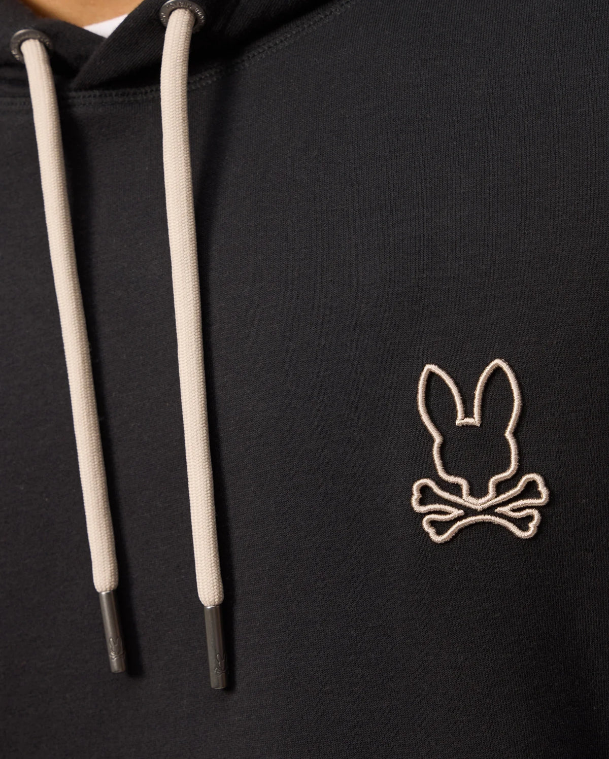 Close-up of a Psycho Bunny MENS JAMES PREMIUM HOODIE - B6H789D200 in black, featuring cream-colored drawstrings. The hoodie showcases an embroidered bunny logo on the left chest area, depicting a stylized rabbit head above crossed bones, outlined in white stitching.