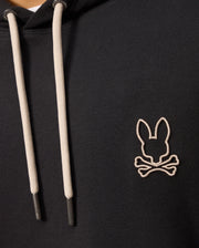Close-up of a Psycho Bunny MENS JAMES PREMIUM HOODIE - B6H789D200 in black, featuring cream-colored drawstrings. The hoodie showcases an embroidered bunny logo on the left chest area, depicting a stylized rabbit head above crossed bones, outlined in white stitching.