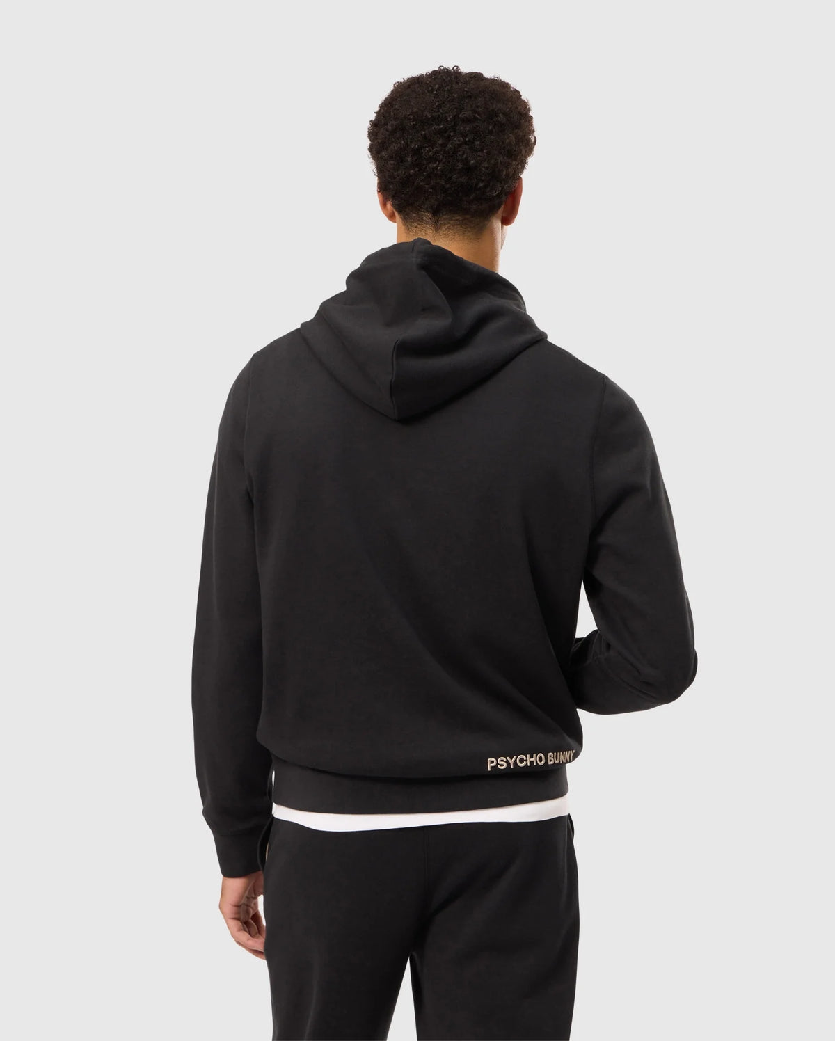 A person with short curly hair is facing away from the camera, wearing a Psycho Bunny MENS JAMES PREMIUM HOODIE - B6H789D200 in black along with black pants. The hoodie features the text "PSYCHO BUNNY" in small letters near the waistline and an embroidered bunny on the back. The background is plain and light-colored.