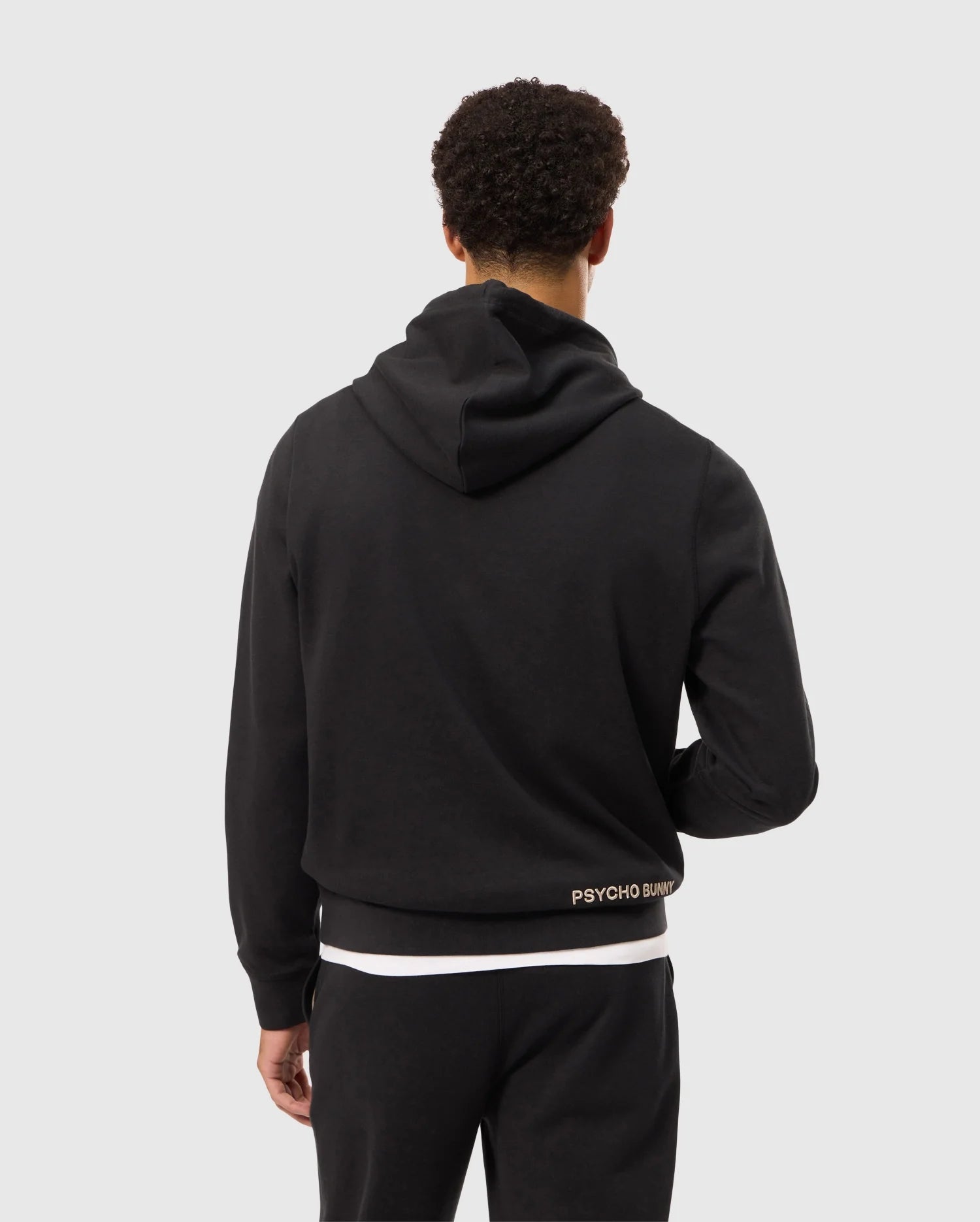 A person with short curly hair is facing away from the camera, wearing a Psycho Bunny MENS JAMES PREMIUM HOODIE - B6H789D200 in black along with black pants. The hoodie features the text "PSYCHO BUNNY" in small letters near the waistline and an embroidered bunny on the back. The background is plain and light-colored.