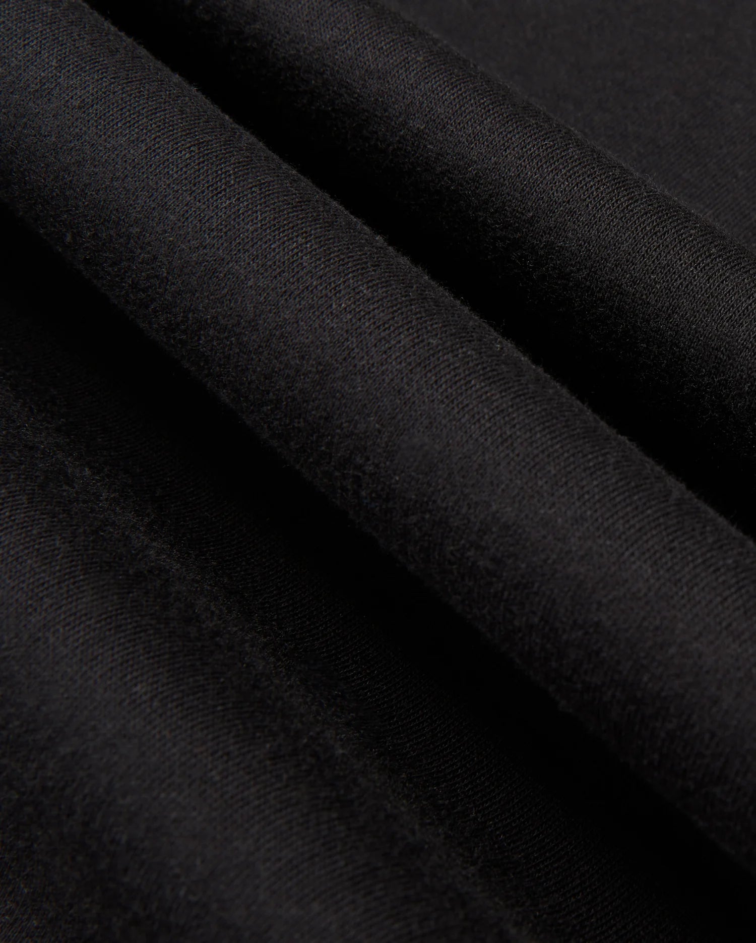 A close-up view of the smooth, black fabric from the Psycho Bunny MENS JAMES PREMIUM HOODIE - B6H789D200 reveals three parallel folds running diagonally across the image. The texture appears soft and even, resembling essential fleece, with a matte finish that creates a subtle interplay of light and shadow on the surface.