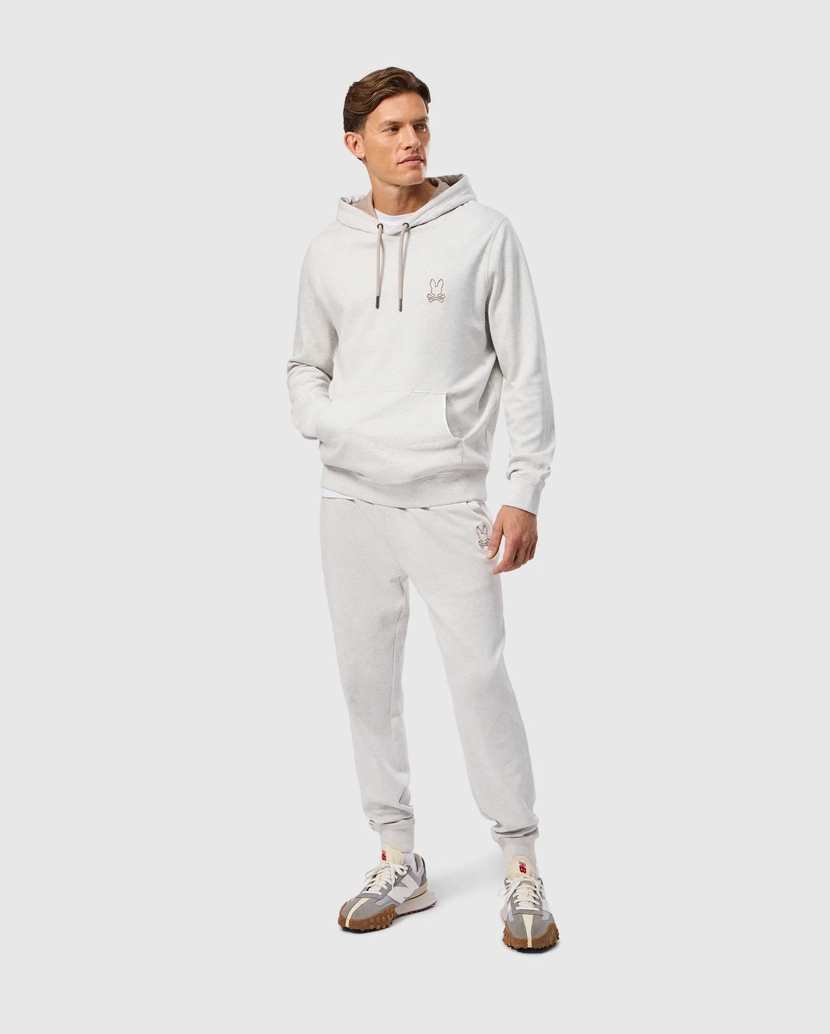 A person wearing the Psycho Bunny MENS JAMES PREMIUM HOODIE (B6H789D200) in light gray, along with matching sweatpants made from high-quality jersey, stands with hands in pockets. They pair the outfit with white and brown sneakers, while a small embroidered bunny logo adorns the chest. The background is a plain light gray.