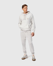 A person wearing the Psycho Bunny MENS JAMES PREMIUM HOODIE (B6H789D200) in light gray, along with matching sweatpants made from high-quality jersey, stands with hands in pockets. They pair the outfit with white and brown sneakers, while a small embroidered bunny logo adorns the chest. The background is a plain light gray.