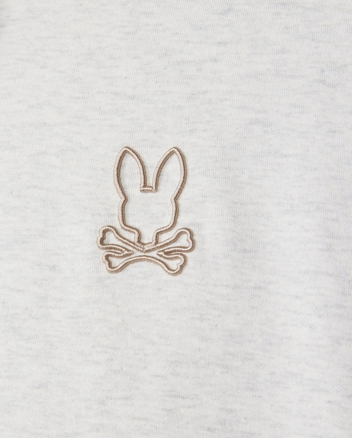 Highlighted in a detailed close-up, the MENS JAMES PREMIUM HOODIE (B6H789D200) from Psycho Bunny features an embroidered bunny design with a rabbit's head outline above two crossed bones. Beige thread contrasts subtly against the gray jersey fabric, infusing stylish warmth into this premium piece.