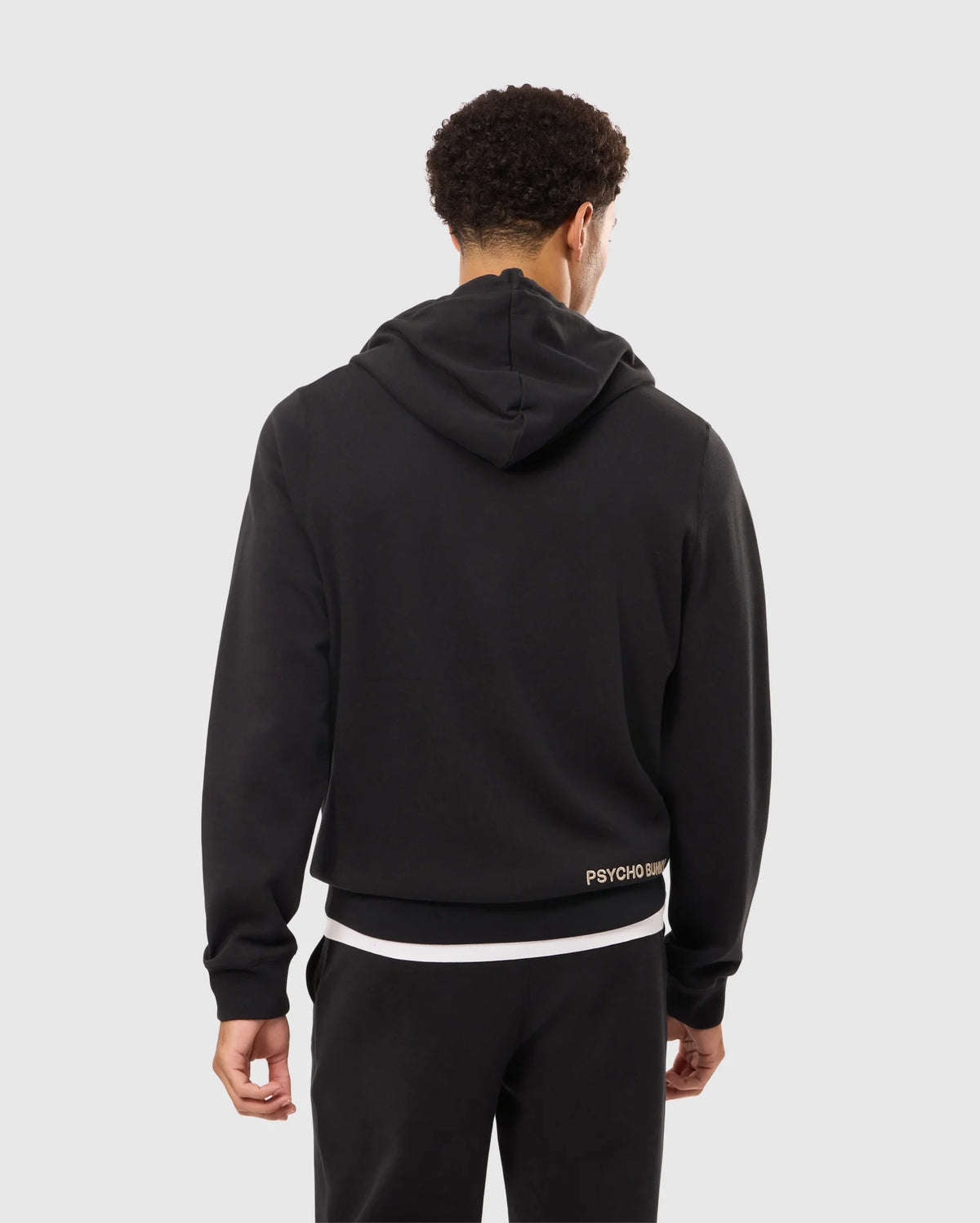 A person with short, curly hair stands facing away from the camera, wearing a black MENS JAMES PREMIUM ZIP HOODIE - B6H793D200 by Psycho Bunny with the hood down and black pants. The back of the hoodie features an embroidered Bunny graphic above the word "PSYCHO" near the bottom right hem.
