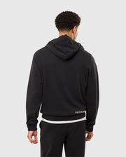 A person with short, curly hair stands facing away from the camera, wearing a black MENS JAMES PREMIUM ZIP HOODIE - B6H793D200 by Psycho Bunny with the hood down and black pants. The back of the hoodie features an embroidered Bunny graphic above the word "PSYCHO" near the bottom right hem.