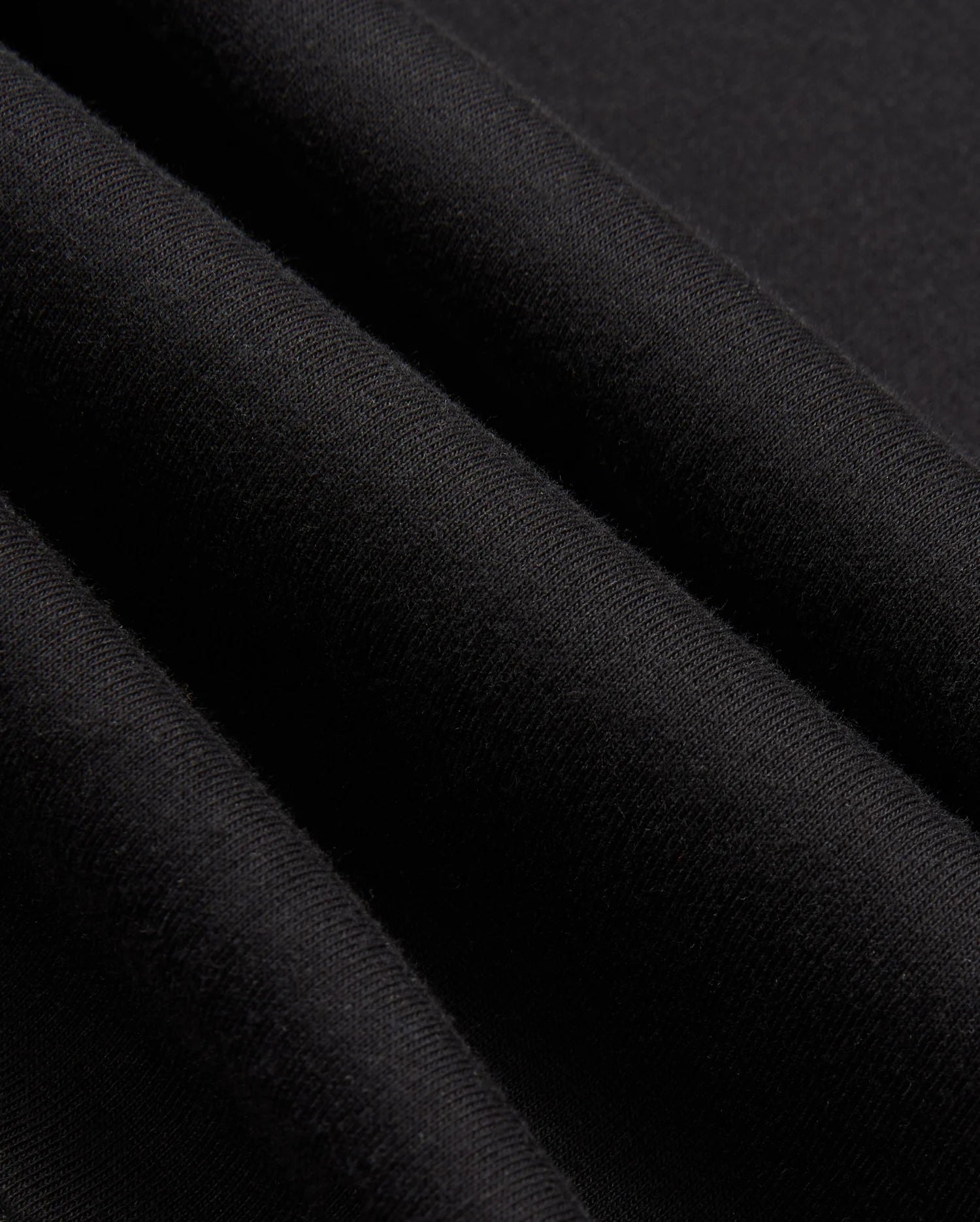 Close-up of the black fabric from the MENS JAMES PREMIUM ZIP HOODIE - B6H793D200 by Psycho Bunny showing its texture. The fabric appears soft and slightly folded, creating gentle shadows and highlights. The material, part of a double-faced premium jersey, looks smooth and uniform in color.
