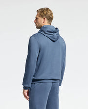 A person wearing a blue Psycho Bunny MENS LEVY ESSENTIAL FLEECE HOODIE - B6H859D200 and matching pants stands facing away against a plain background, showcasing a side profile. Crafted from 100% cotton French terry, the ensemble complements the individual's short, light-colored hair.