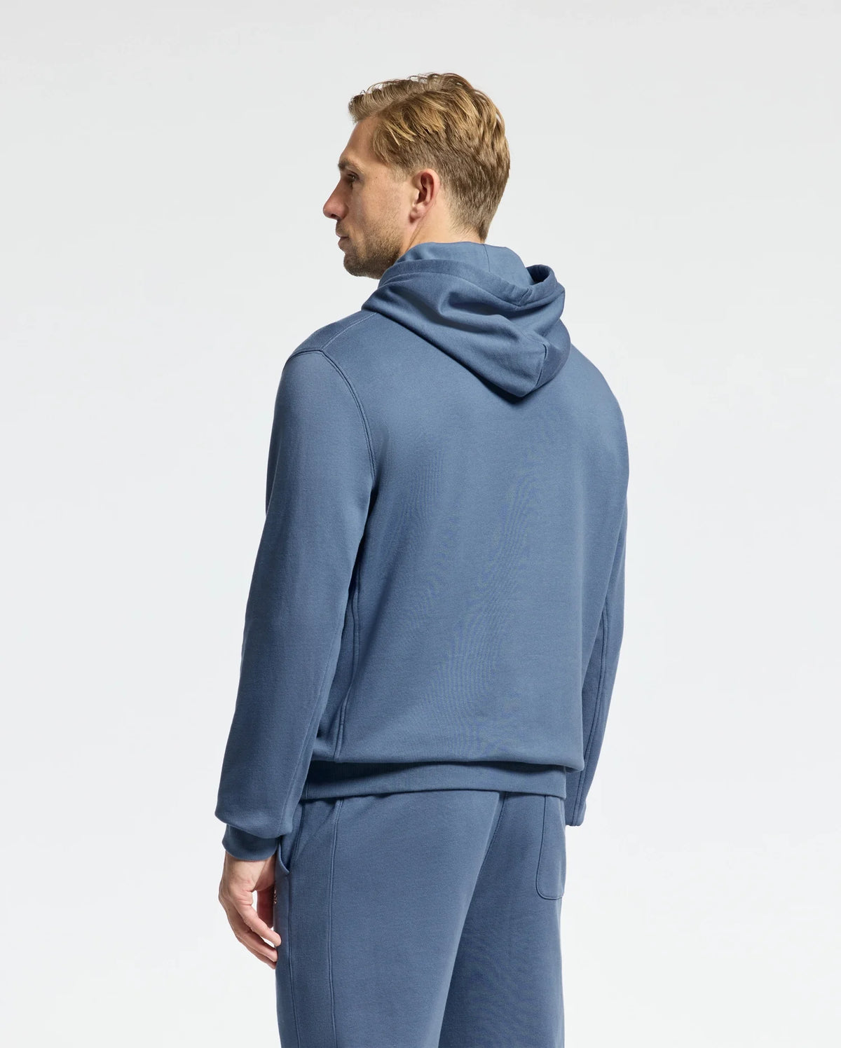 A person wearing a blue Psycho Bunny MENS LEVY ESSENTIAL FLEECE HOODIE - B6H859D200 and matching pants stands facing away against a plain background, showcasing a side profile. Crafted from 100% cotton French terry, the ensemble complements the individual's short, light-colored hair.