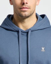 A man is seen wearing the blue MENS LEVY ESSENTIAL FLEECE HOODIE - B6H859D200 by Psycho Bunny, constructed from soft French terry and adorned with a small white bunny and crossbones logo on the chest. The image shows the lower half of his face and upper chest against a simple background.