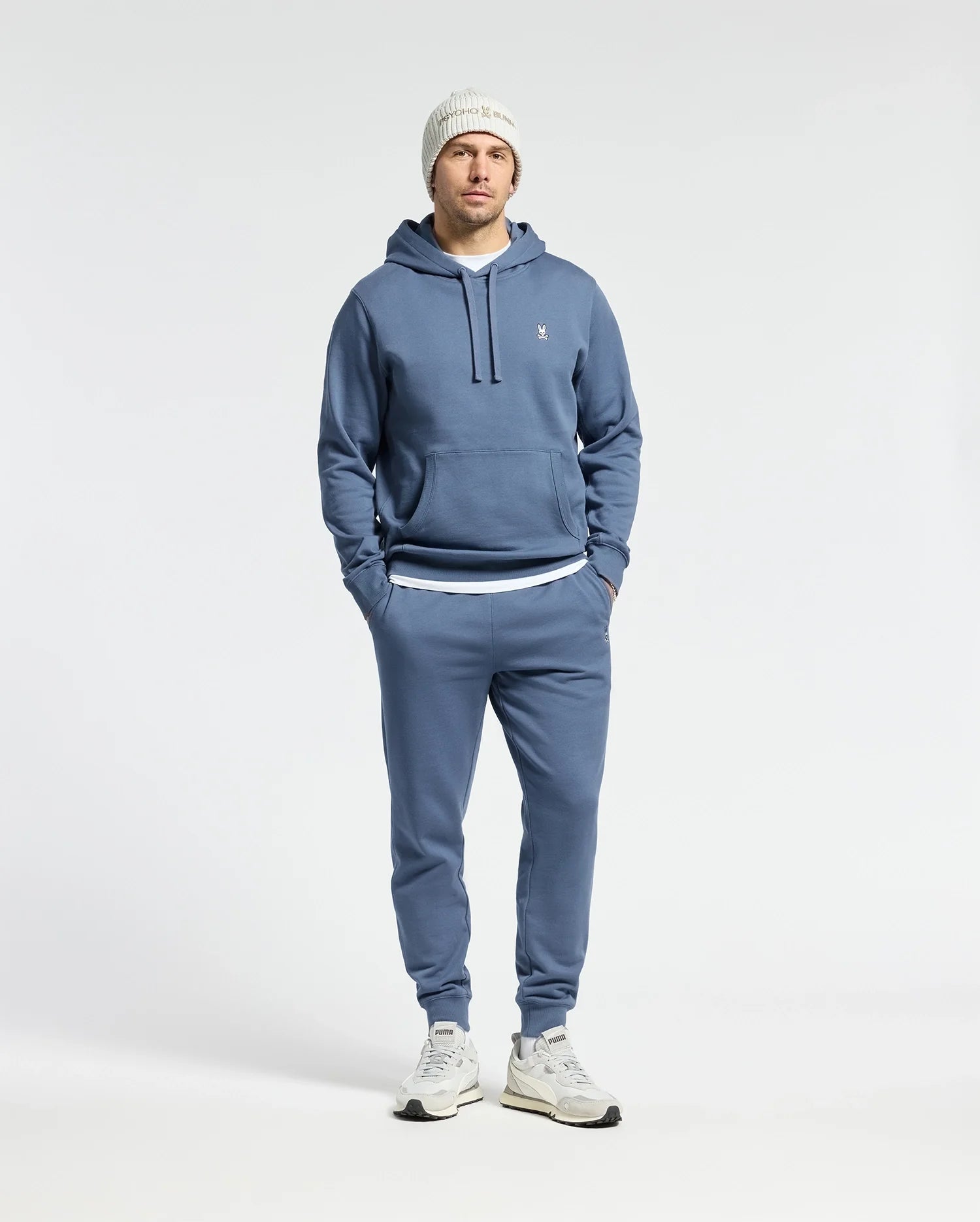 A man stands against a white background wearing the Psycho Bunny MENS LEVY ESSENTIAL FLEECE HOODIE - B6H859D200 in blue, paired with matching joggers, a white knit beanie, and 100% cotton white sneakers. His hands are in the hoodie pockets.