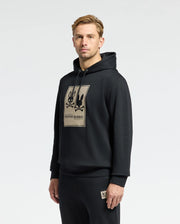 A person wearing the comfortable MENS MONROE TWILL PATCH FLEECE HOODIE - B6H926D200, styled in black and featuring the distinctive "Psycho Bunny" logo, stands against a plain white background. This hoodie, focused on comfort, boasts a branded patch on the chest. The individual, with short hair, gazes directly at the camera.