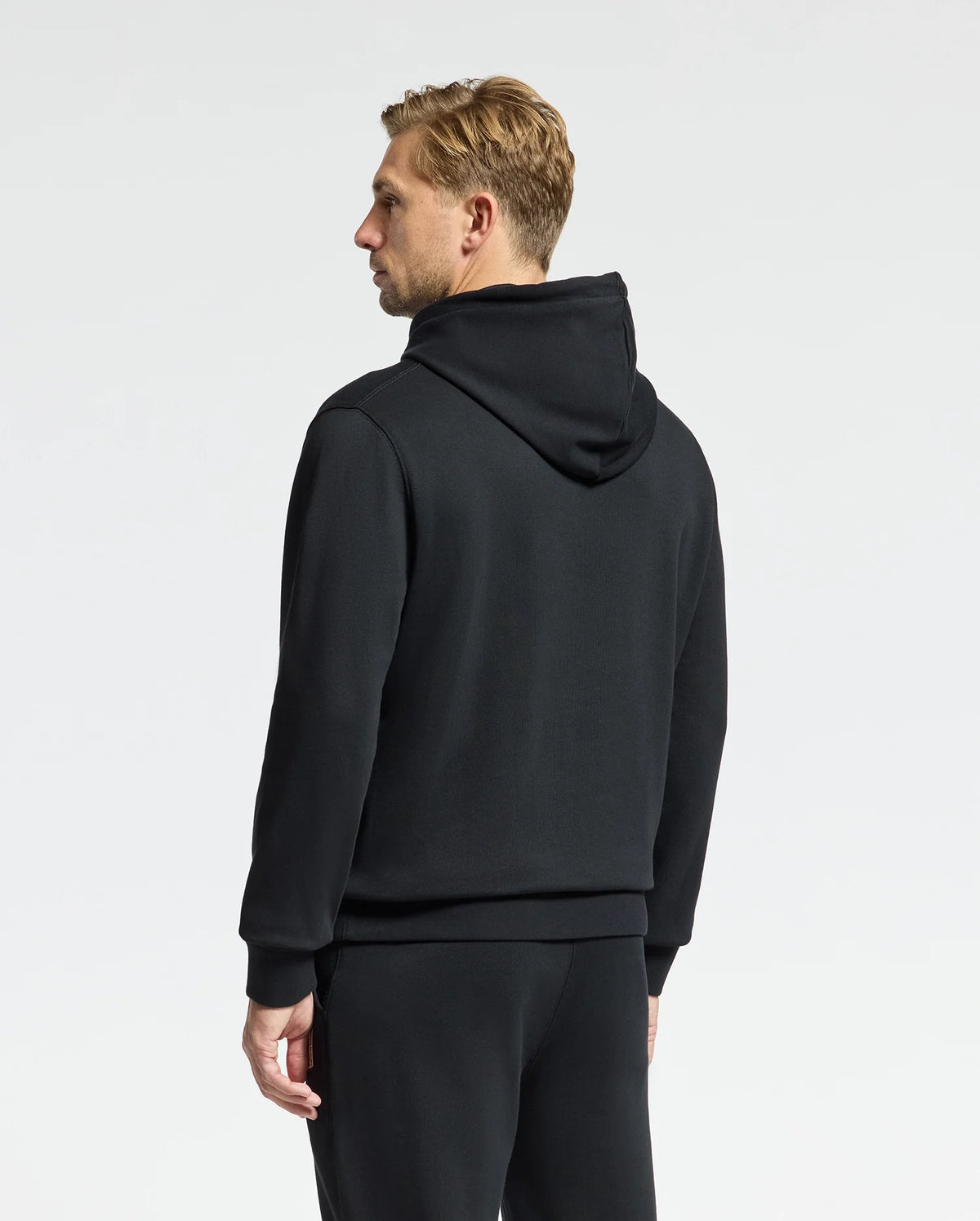 Against a plain background, an individual stands dressed in a black MENS MONROE TWILL PATCH FLEECE HOODIE from Psycho Bunny, paired with cozy sweatpants. They face away from the camera as their head slightly turns to the side, showing their short, light hair.