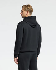Against a plain background, an individual stands dressed in a black MENS MONROE TWILL PATCH FLEECE HOODIE from Psycho Bunny, paired with cozy sweatpants. They face away from the camera as their head slightly turns to the side, showing their short, light hair.