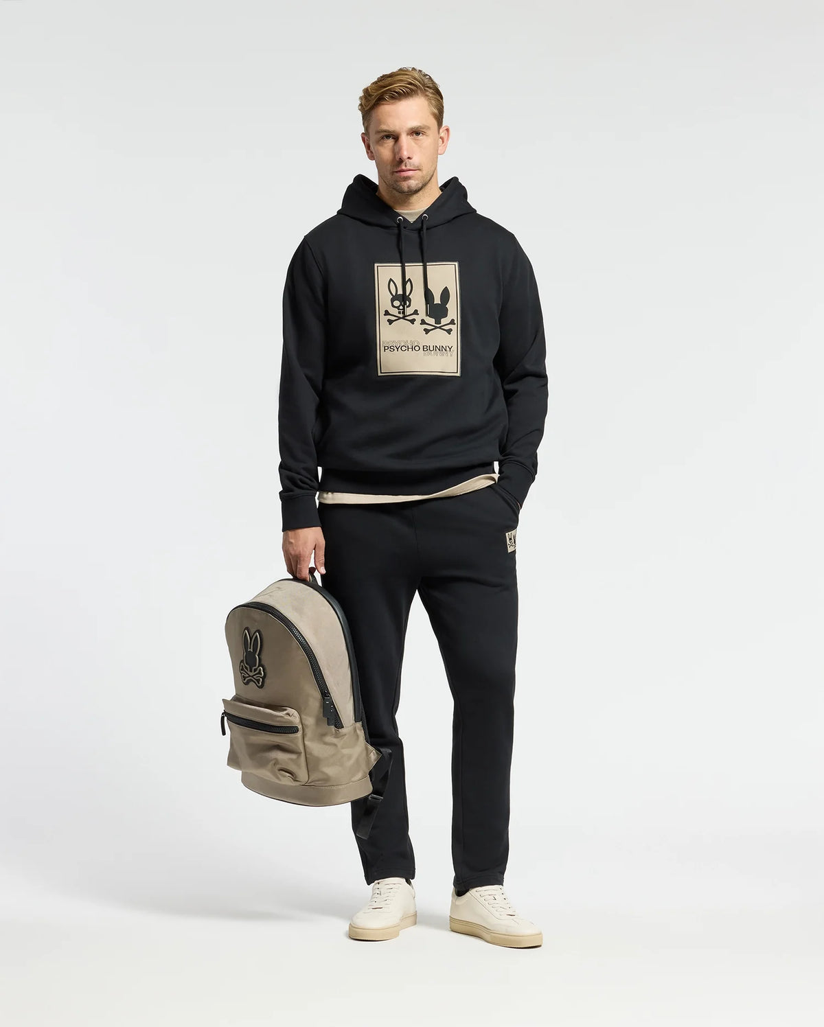 A man in a Psycho Bunny MONROE twill patch fleece hoodie with a graphic design stands holding a beige backpack. He wears matching black sweatpants and beige sneakers, blending style and comfort against a plain white background.