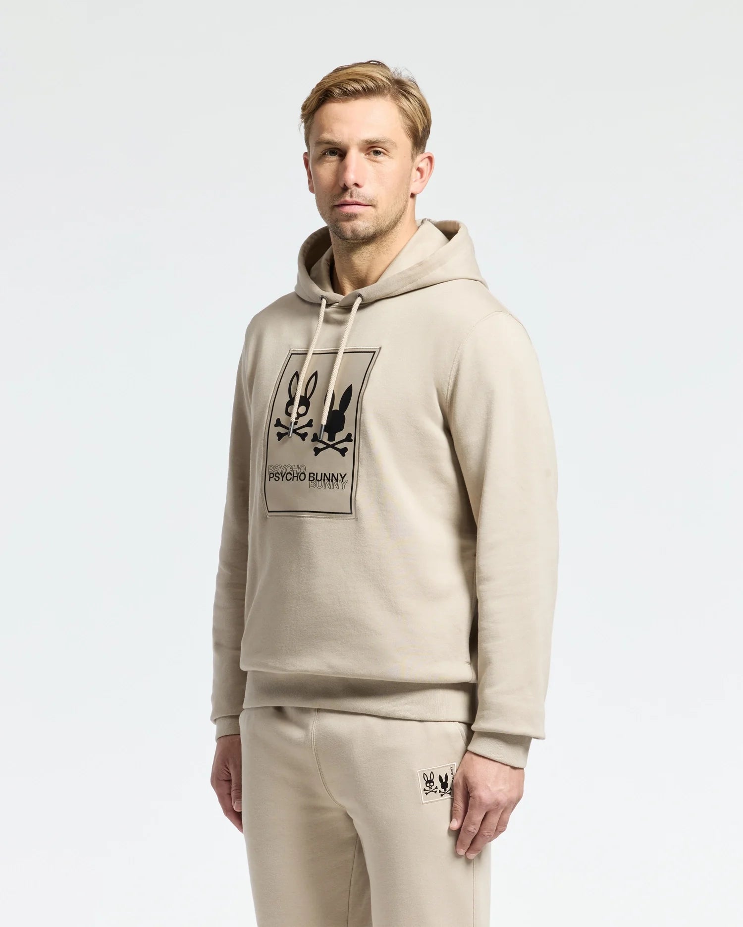 A man is wearing a Psycho Bunny MENS MONROE TWILL PATCH FLEECE HOODIE in beige, along with matching sweatpants, both featuring the brand's logo appliqué. He stands against a plain white background, looking at the camera with a neutral expression.