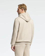 A man in a Psycho Bunny MENS MONROE TWILL PATCH FLEECE HOODIE, beige in color, paired with matching pants, stands with his back to the camera against a neutral background.