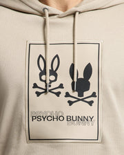 Close-up of the Psycho Bunny Men's Monroe Twill Patch Fleece Hoodie - B6H926D200 in beige, crafted from soft French terry. It features the iconic Psycho Bunny logo appliqué with two stylized bunny skulls and crossbones, along with the bold black text "PSYCHO" and "BUNNY" below the images.