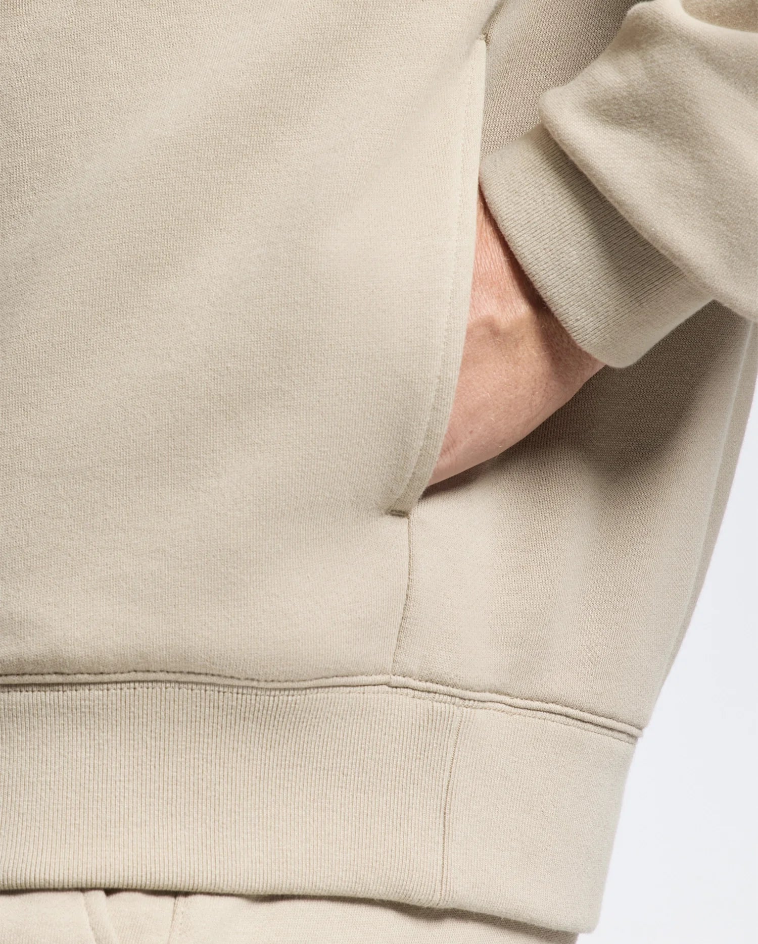 Close-up of a person wearing the beige Psycho Bunny Men's Monroe Twill Patch Fleece Hoodie (B6H926D200), with their hand tucked into a side pocket. The fabric boasts a soft, fleece texture. The image focuses on the midsection, highlighting the garment's design and texture.