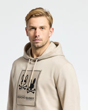 A man wearing a Psycho Bunny MENS MONROE TWILL PATCH FLEECE HOODIE - B6H926D200, adorned with a black graphic of a bunny and crossbones design, features the iconic "Psycho Bunny" logo appliqué, standing out against the plain white background.