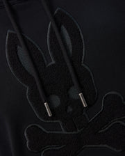 Close-up of the Psycho Bunny MENS PACIFIC CHENILLE HOODIE - B6H930D200 in black, highlighting chenille embroidery featuring a stylized skull with bunny ears atop crossed bones. The cotton knit strings are visible and feature metal-capped tips.