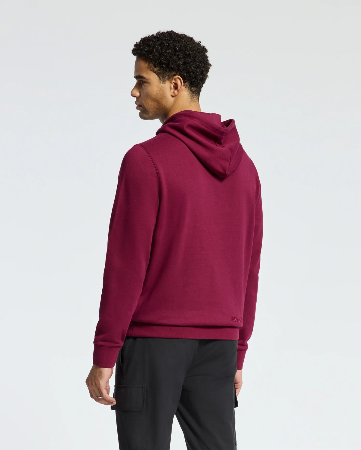 A man stands facing sideways against a neutral background, wearing the MENS PACIFIC HIGH PILE CHENILLE HOODIE - B6H930D200 by Psycho Bunny in maroon and paired with black pants. His hands rest by his sides as he looks slightly downward.