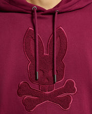 Close-up of the Psycho Bunny MENS PACIFIC HIGH PILE CHENILLE HOODIE - B6H930D200 in luxurious maroon cotton, featuring an embroidered rabbit skull and crossbones on the chest. The hoodie strings are visible above the design, enhancing its stylish allure.