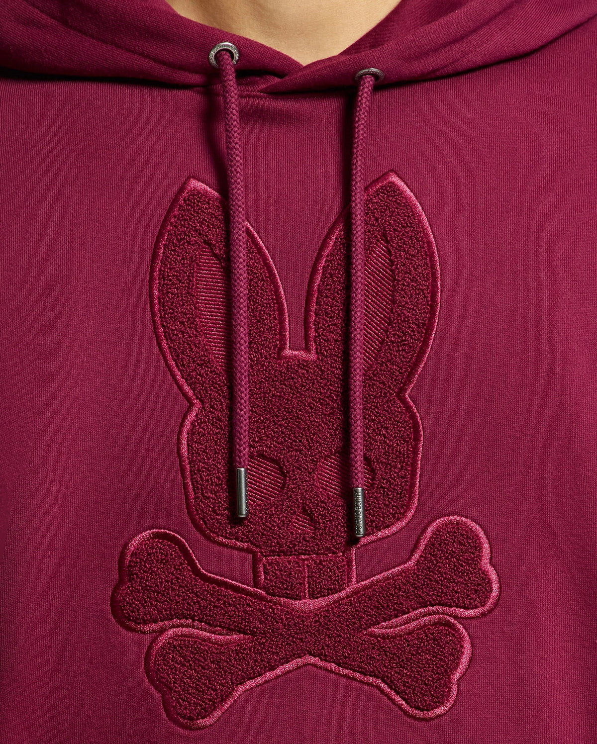 Close-up of the Psycho Bunny MENS PACIFIC HIGH PILE CHENILLE HOODIE - B6H930D200 in luxurious maroon cotton, featuring an embroidered rabbit skull and crossbones on the chest. The hoodie strings are visible above the design, enhancing its stylish allure.