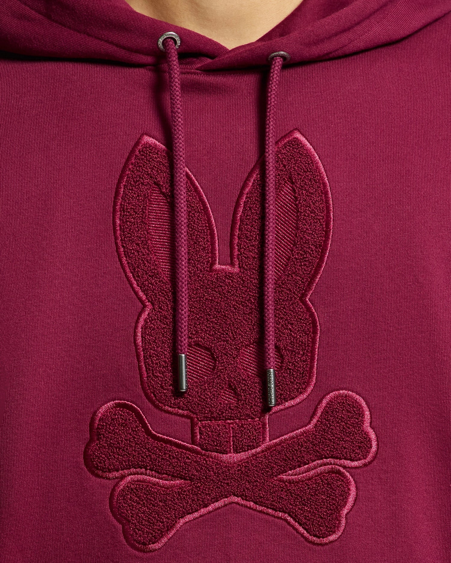 Close-up of the Psycho Bunny MENS PACIFIC HIGH PILE CHENILLE HOODIE - B6H930D200 in luxurious maroon cotton, featuring an embroidered rabbit skull and crossbones on the chest. The hoodie strings are visible above the design, enhancing its stylish allure.