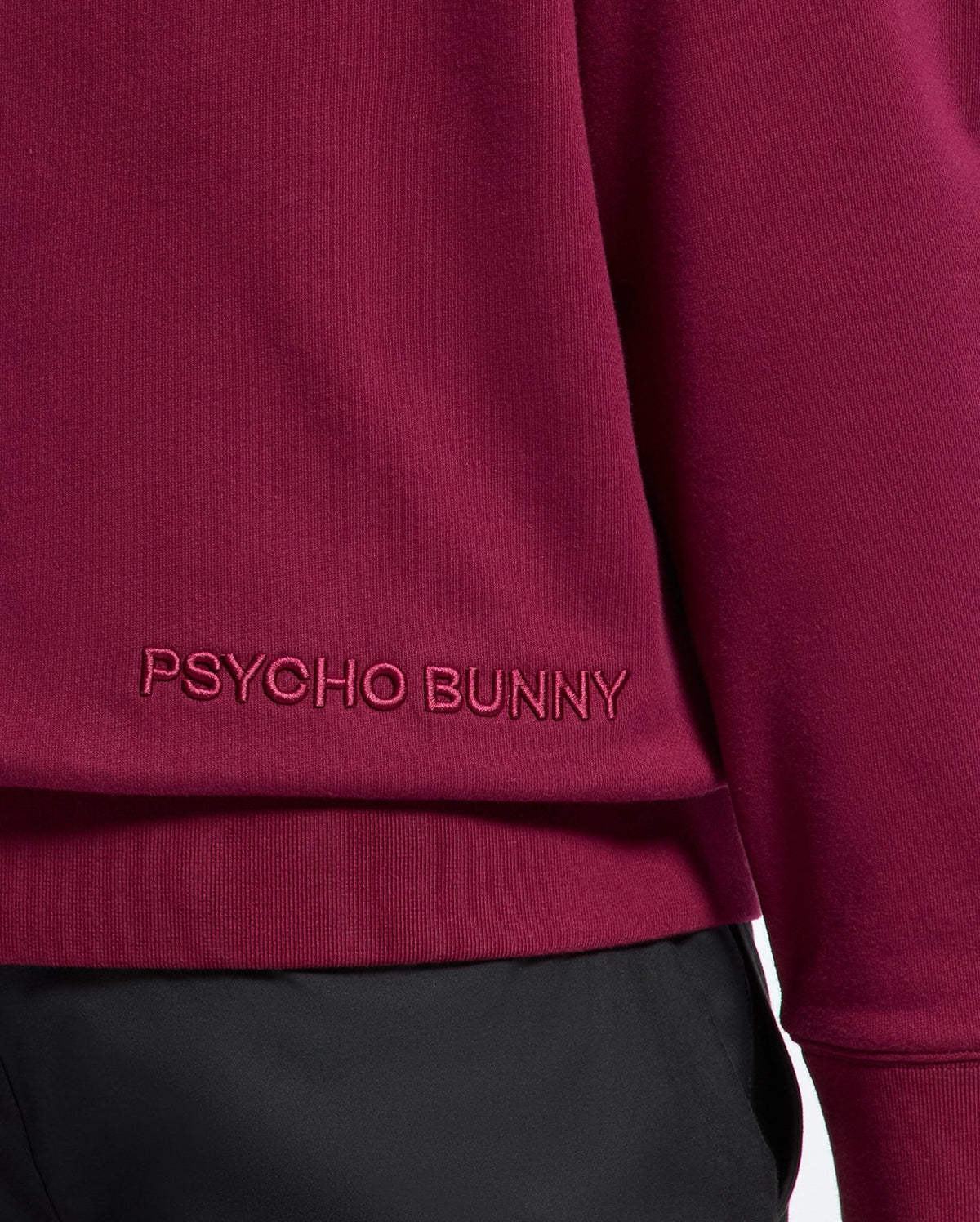 Close-up of a maroon luxurious cotton hoodie, branded as the "MENS PACIFIC HIGH PILE CHENILLE HOODIE - B6H930D200" by Psycho Bunny, with their name embroidered near the hem. It is paired with black pants.
