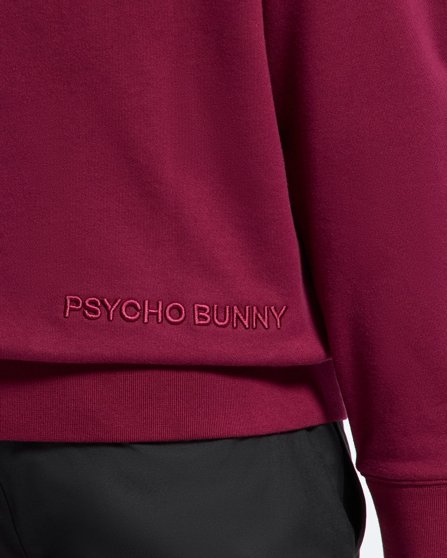 Close-up of a maroon luxurious cotton hoodie, branded as the "MENS PACIFIC HIGH PILE CHENILLE HOODIE - B6H930D200" by Psycho Bunny, with their name embroidered near the hem. It is paired with black pants.