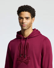 A person wearing a maroon Psycho Bunny MENS PACIFIC HIGH PILE CHENILLE HOODIE - B6H930D200, adorned with a bunny and crossbones design, stands against a plain background. They have short, curly hair and a neutral expression.