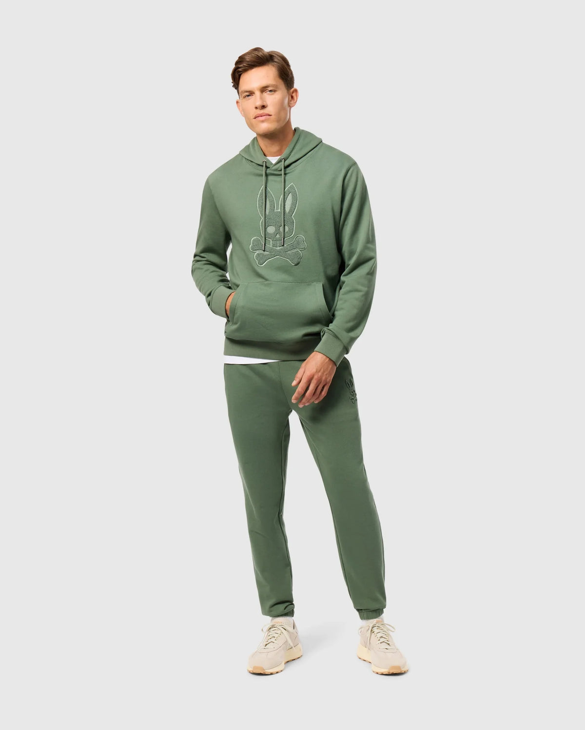 A person is dressed in the MENS PACIFIC CHENILLE HOODIE - B6H930D200 from Psycho Bunny, made of soft French terry fabric, paired with matching sweatpants. The ensemble features chenille embroidery with a cartoon graphic. They're wearing beige sneakers and have one hand in their pocket while standing against a plain, light-colored background.