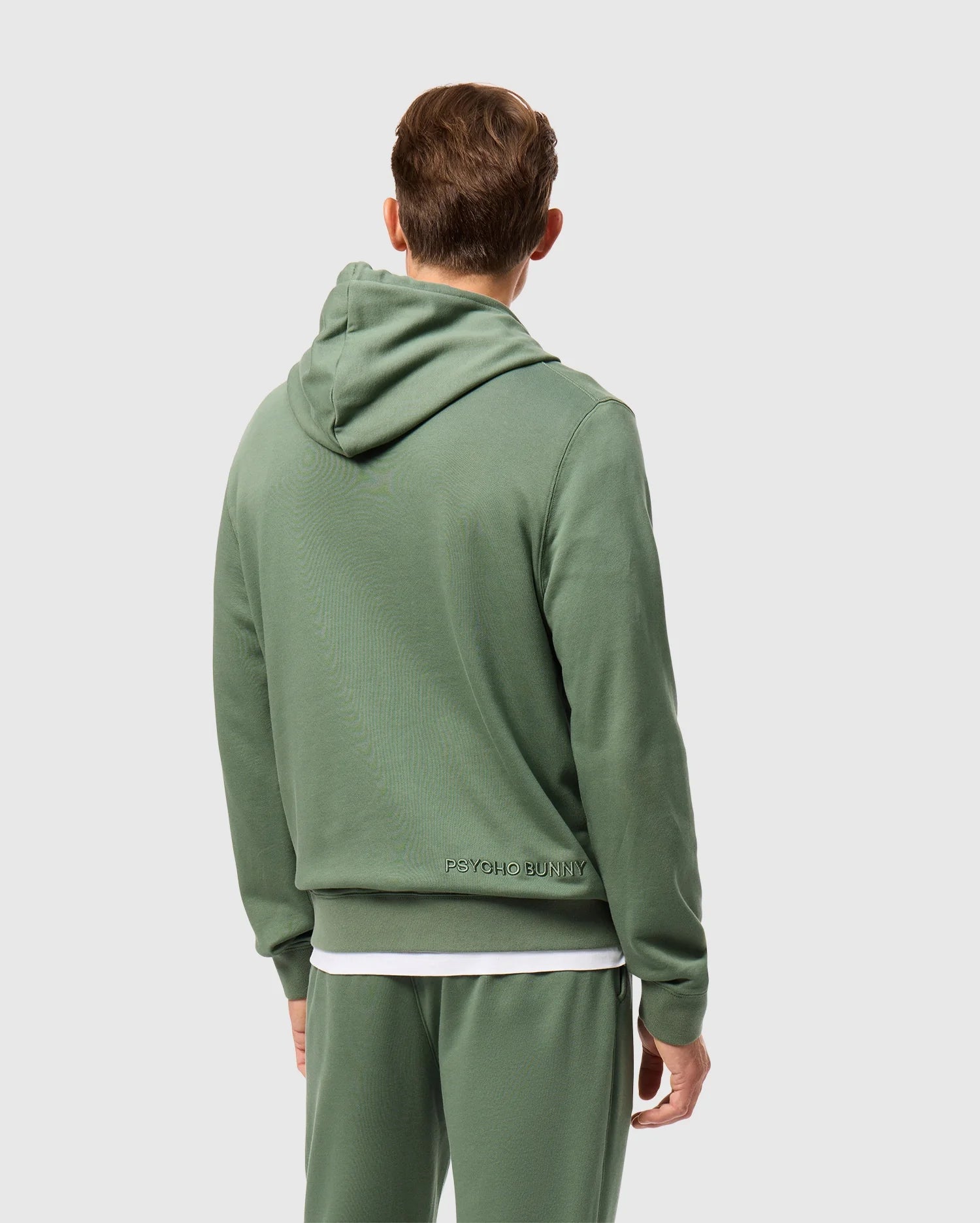 A person wearing a green Psycho Bunny hoodie, specifically the Men's Pacific Chenille Hoodie (B6H930D200), paired with matching sweatpants is seen from behind. The French terry hoodie showcases "Psycho Bunny" in chenille embroidery on the lower back. The individual, who has short brown hair, stands against a light gray background.