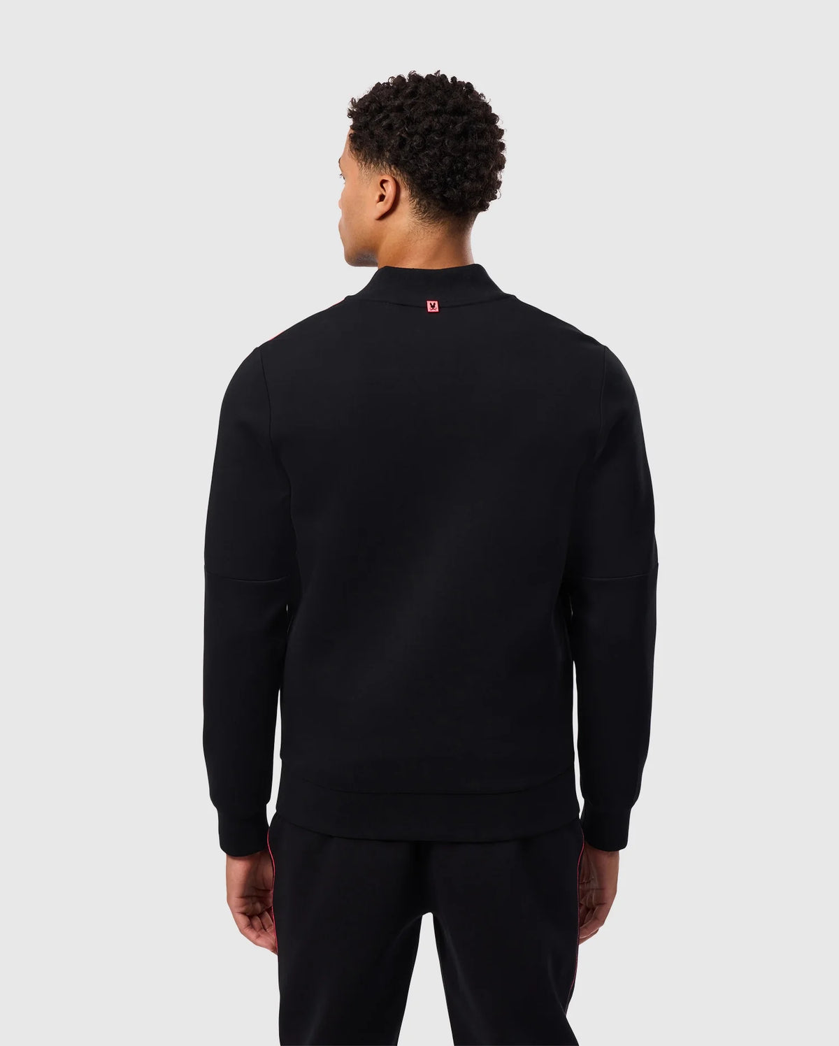 A person with short curly hair, seen from the back, is wearing the Psycho Bunny MENS TRAVIS TRACK JACKET - B6J142D200 in black paired with black pants. The background is light gray.