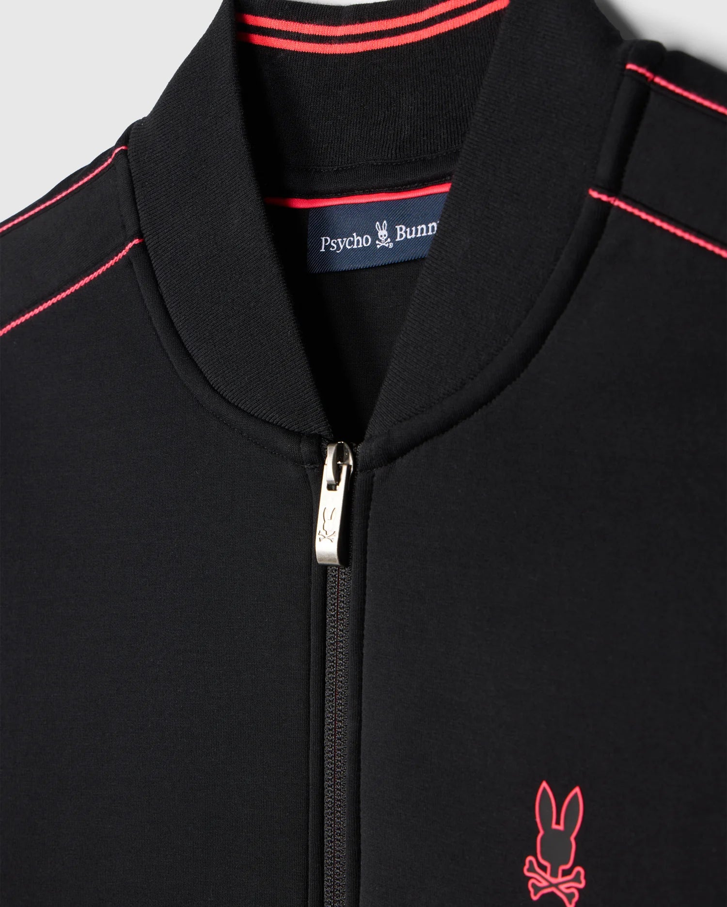 Close-up of the Psycho Bunny MENS TRAVIS TRACK JACKET - B6J142D200 in black with red detailing. The collar and shoulders feature thin red stripes, and a small embroidered bunny logo with crossbones appears on the left side of the chest. Invisible zip pockets add functionality to this stylish piece.