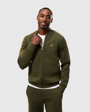 A person is smiling at the camera while wearing a high-quality cotton-blend olive green Psycho Bunny MENS TRAVIS TRACK JACKET (B6J142D200) with orange piping, part of a matching set. The tracksuit top is zipped up halfway, revealing a white shirt underneath. They have one hand in their pocket and are posing against a plain, light grey background.