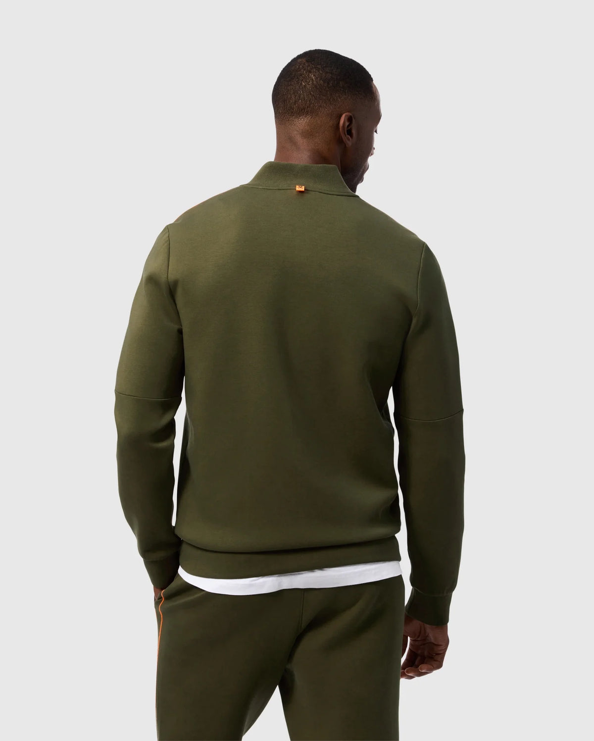 Rear view of a person wearing an olive green Psycho Bunny MENS TRAVIS TRACK JACKET - B6J142D200 and matching pants, standing against a plain white background. The high-quality cotton-blend jacket features a small brown tag on the back of the collar. The person also appears to be wearing a white T-shirt underneath the jacket.
