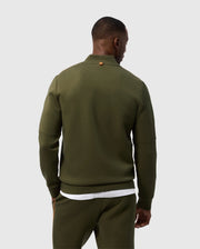 Rear view of a person wearing an olive green Psycho Bunny MENS TRAVIS TRACK JACKET - B6J142D200 and matching pants, standing against a plain white background. The high-quality cotton-blend jacket features a small brown tag on the back of the collar. The person also appears to be wearing a white T-shirt underneath the jacket.