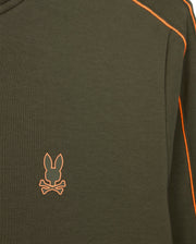 Close-up of the Psycho Bunny MENS TRAVIS TRACK JACKET - B6J142D200, a dark green high-quality cotton-blend garment with orange stitching along the seams. This jacket, part of a matching set, features an orange emblem on the left chest area depicting a rabbit head above crossed bones.