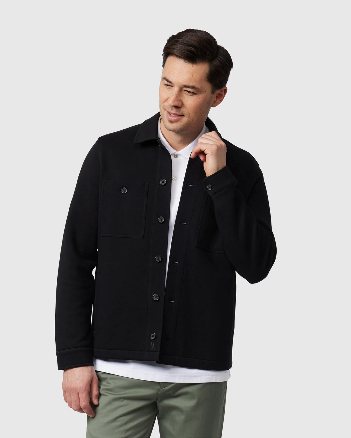 A person wearing a black Psycho Bunny MENS BEDFORD SHIRT JACKET - B6J290B200 over a white shirt and green pants stands against a plain background, looking down with one hand by their face.