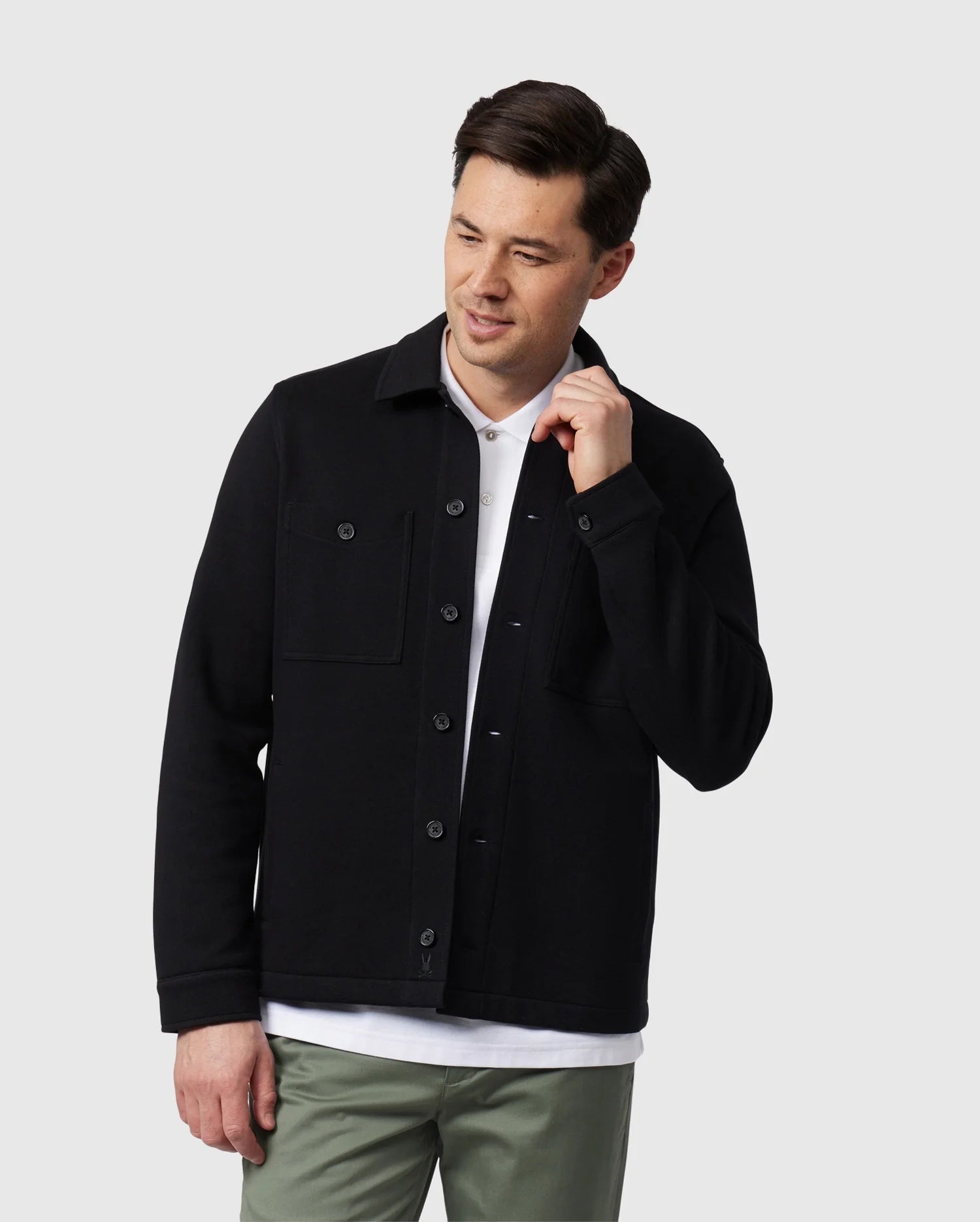 A person wearing a black Psycho Bunny MENS BEDFORD SHIRT JACKET - B6J290B200 over a white shirt and green pants stands against a plain background, looking down with one hand by their face.