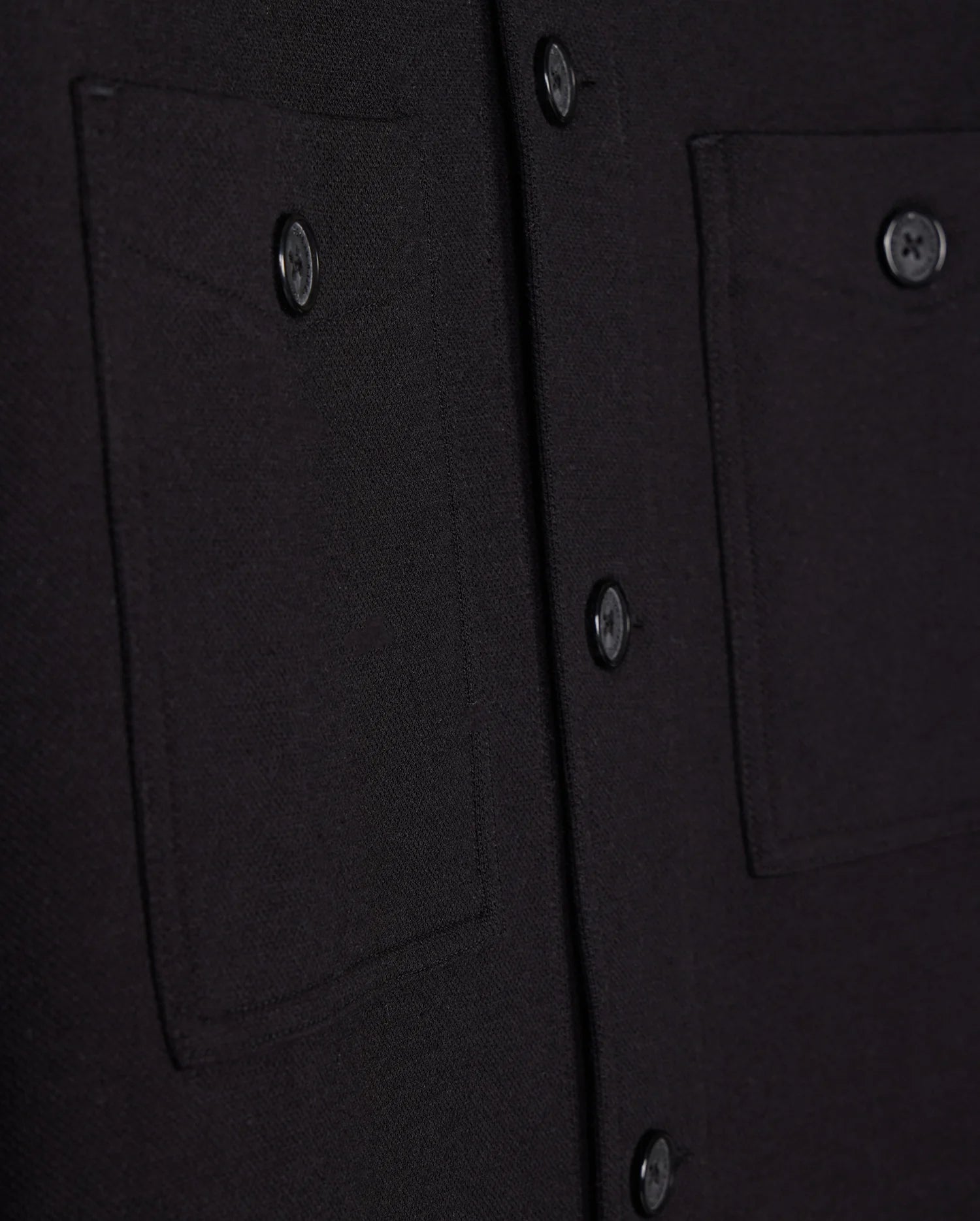 Close-up of the Psycho Bunny Men's Bedford Shirt Jacket - B6J290B200 in black fabric, showcasing two pockets and a matching row of buttons. The textured material evokes a work-jacket style, with pockets featuring button closures.