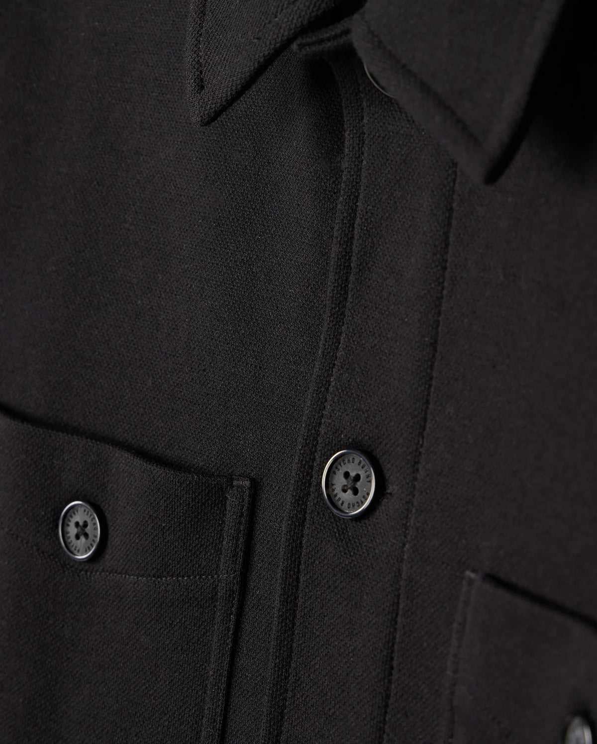 Close-up of the Psycho Bunny MENS BEDFORD SHIRT JACKET - B6J290B200 in black, showcasing two dark buttons and dual chest pockets. The fabric exhibits a textured appearance analogous to work-jacket style, with the men's jacket neatly buttoned.