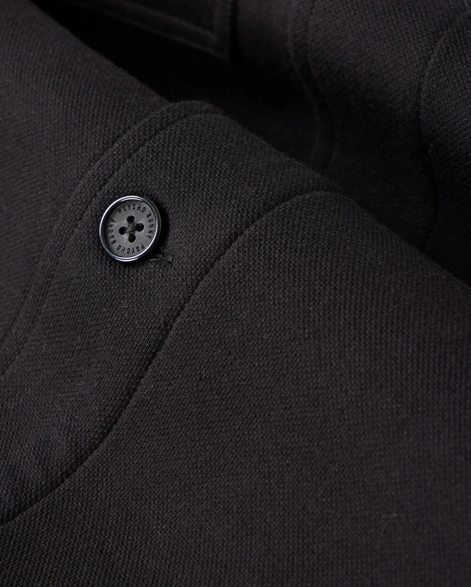 Zoomed-in view of the black fabric from the MENS BEDFORD SHIRT JACKET - B6J290B200 by Psycho Bunny, highlighting the texture and stitching details with a single black button, reminiscent of a work-jacket style.