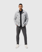 A man confidently poses against a plain background, wearing the Psycho Bunny BEDFORD SHIRT JACKET - B6J290B200 in light gray over a black shirt, paired with dark gray pants and white sneakers. His hands are tucked into his jacket pockets as he stands facing forward.