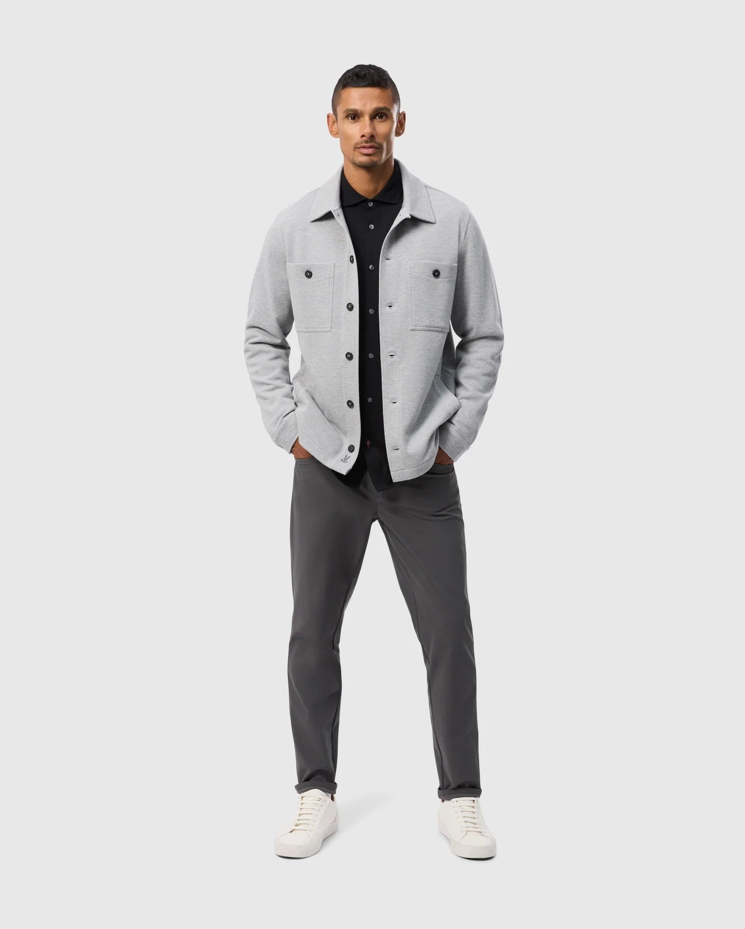 A man confidently poses against a plain background, wearing the Psycho Bunny BEDFORD SHIRT JACKET - B6J290B200 in light gray over a black shirt, paired with dark gray pants and white sneakers. His hands are tucked into his jacket pockets as he stands facing forward.